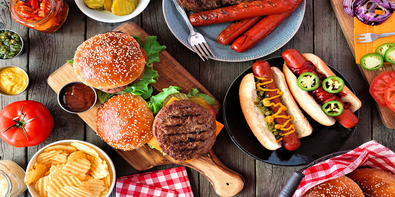 Summer BBQ food table scene with hot dog and hamburger buffet. Top view over a dark wood background.